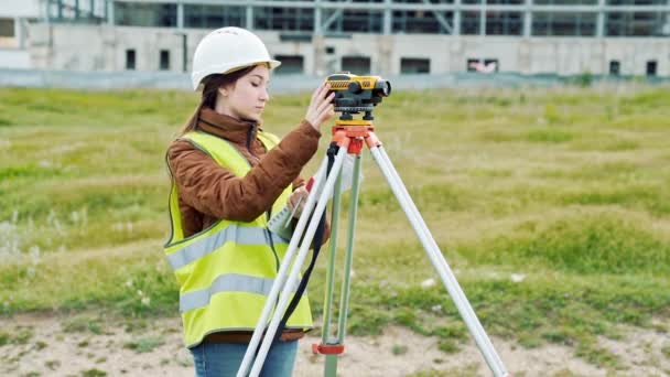 En ung kvinna besiktningsman i arbetskläder och hjälm justerar utrustningen och producerar beräkningar på byggarbetsplatsen. Begreppet landskap design, geodesi — Stockvideo