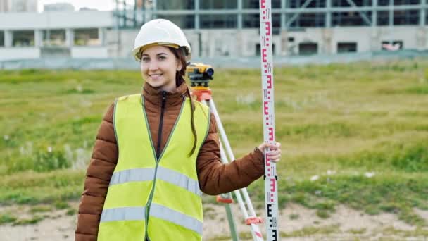 Jonge smileing vrouw landmeter in groene werkkleding en helm past de apparatuur, produceert berekeningen en schrijft in het notitieblok op de bouwplaats. Het concept van het ontwerp van het landschap — Stockvideo