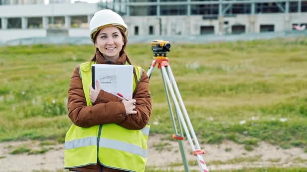 Joven sonriente topógrafa en ropa de trabajo verde y casco ajusta el equipo, produce cálculos y escribe al cuaderno en el sitio de construcción. El concepto de diseño paisajístico — Vídeo de stock