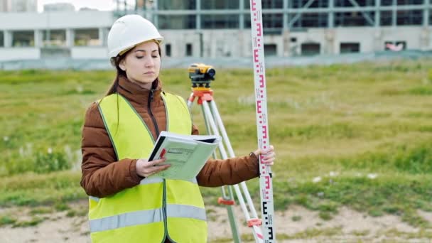 Een jonge vrouw landmeter in werkkleding en helm past de apparatuur en produceert berekeningen op de bouwplaats. Het concept van landschap design, geodesie — Stockvideo