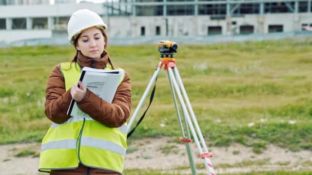 一名年轻女子的工作服和头盔的测量师调整设备, 并在建筑工地上产生计算。景观设计的概念, 大地测量学 — 图库视频影像