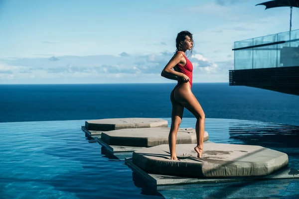 Amazing view of sexy girl relaxing at swimming pool with ocean at background. Attractive young woman in red bikini posing. Concept of swimsuit, swimwear and travel — Stock Photo, Image