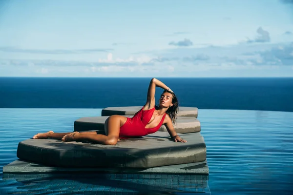Hermosa vista de chica sexy relajarse en la piscina con el océano en el fondo. Preciosa joven en bikini rojo posando. Concepto de traje de baño, traje de baño y viajes — Foto de Stock