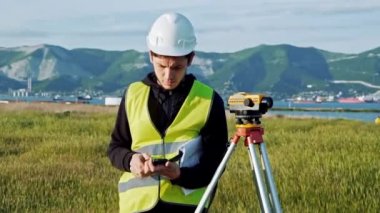 yeşil iş elbiseleri ve kask Surveyor erkekte ekipman ayarlar, hesaplamalar üretir ve inşaat alanının üstünde belgili tanımlık defter yazar. Peyzaj konsept tasarımı, Jeodezi