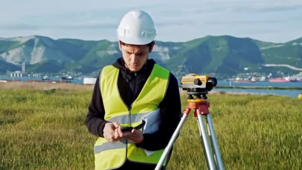 Surveyor man i gröna kläder och hjälm justerar utrustningen, producerar beräkningar och skriver i anteckningsboken på byggarbetsplatsen. Begreppet landskap design, geodesi — Stockvideo