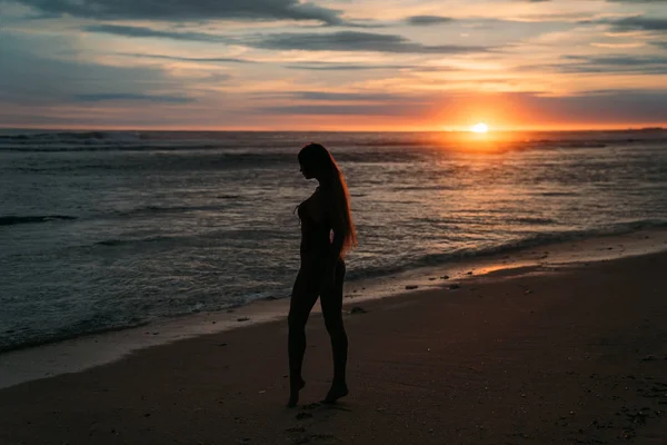 Silhouette einer hübschen Frau, Seitenansicht. schönes junges Mädchen mit langen Haaren, das am Strand spaziert und bei Sonnenuntergang posiert. Konzept des Reisens, der Entspannung, des Yoga — Stockfoto