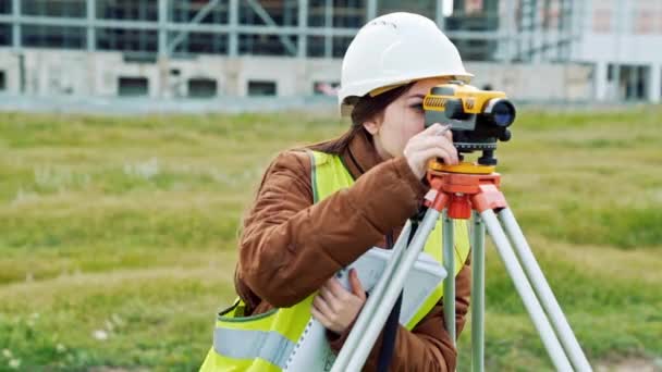 En ung kvinna besiktningsman i arbetskläder och hjälm justerar utrustningen och producerar beräkningar på byggarbetsplatsen. Begreppet landskap design, geodesi — Stockvideo