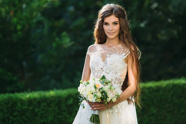 Bonito jovem noiva com cabelos longos segurando seu buquê de casamento inclui rosas brancas e outras flores. Vestido de casamento branco bonito. Menina bonita em árvores cumprimentar fundo — Fotografia de Stock