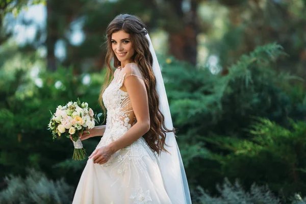 彼女の結婚式のブーケを保持している長い毛でかわいい若い花嫁には、白いばらおよび他の花が含まれています。美しい白結婚ドレス。きれいな女の子を迎える木バック グラウンド — ストック写真