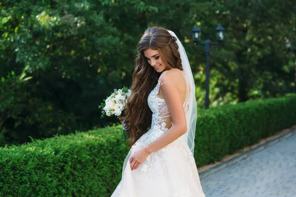 Porträtt av vackra unga bruden med söt smite i vit bröllop klänning håll bukett blommor i hennes händer står på grönt gräs bakgrund. Begreppet kläder och familj — Stockfoto