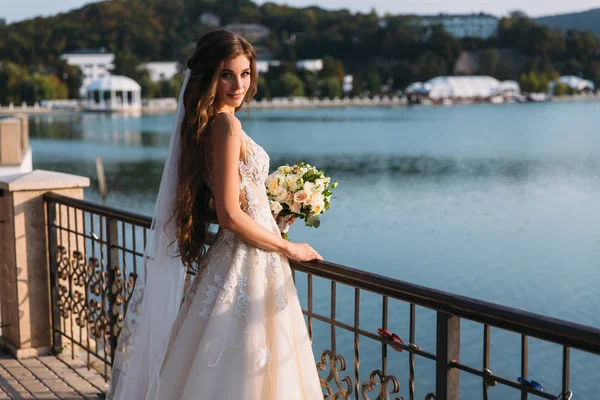 Mooie jonge bruid in witte trouwjurk met lange haren, boeket bloemen in haar handen staat, meer op de achtergrond. — Stockfoto