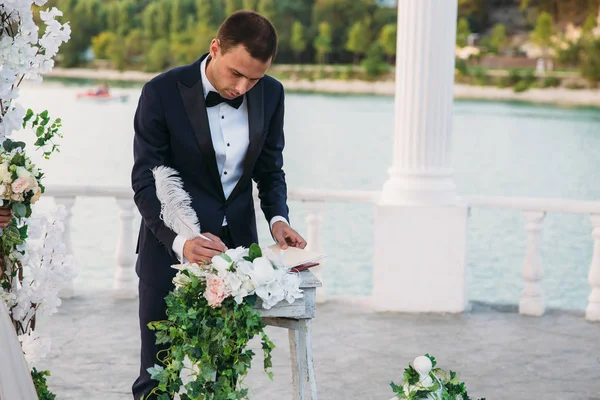 黒のジャケットと結婚式の日に蝶ネクタイと白いシャツで新郎に背景に湖のあるアーチの近く署名を入れてください。 — ストック写真