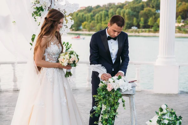 黒のジャケットと結婚式の日に蝶ネクタイと白いシャツで、新郎は、背景に湖のあるアーチの近くの署名を置きます。新婚夫婦が一緒に立っています。花のブーケと花嫁 — ストック写真