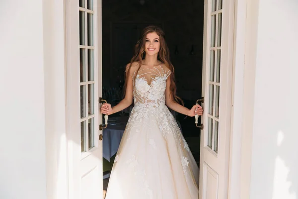 Schöne Braut mit attraktiven langen Haaren steht neben weißen Türen. hübsche junge Frau im weißen Kleid mit Hochzeitsschminke. Nahaufnahme Porträt lächelnde Braut — Stockfoto