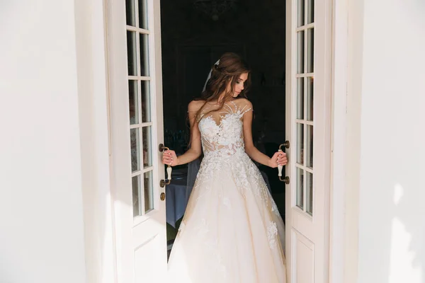 Vacker brud med vackra långa hårstrån står nära vita dörrar. Ganska ung kvinna i vit klänning med bröllop make-up. Närbild porträtt leende bruden — Stockfoto