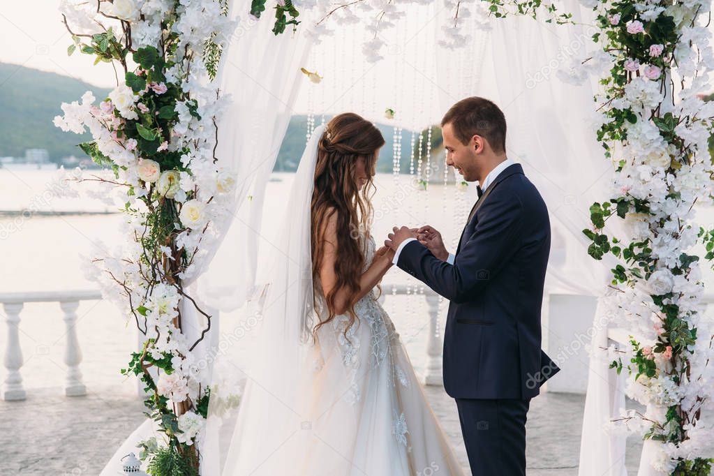 couple in wedding arch exchange rings with lake on background, the bride with long beautiful hairs and groom in black suit look at each other in wedding day. Concept of love and family