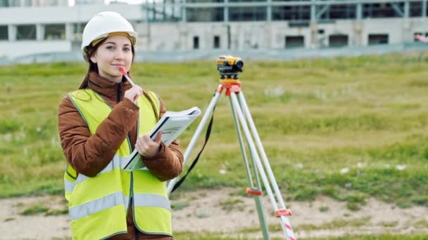 Jeune femme souriante arpenteur en vert vêtements de travail et casque ajuste l'équipement, produit des calculs et écrit sur le carnet sur le chantier de construction. Le concept d'aménagement paysager — Video