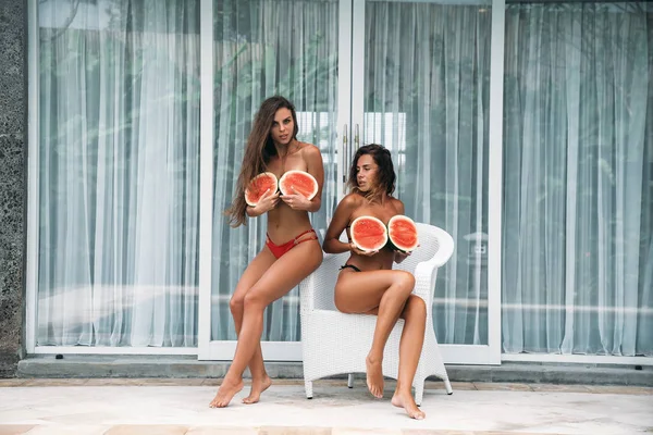 Retrato de dos morenas en traje de baño con fruta de sandía sosteniendo en las manos y cubrir el pecho. Modelo sexy con cuerpo perfecto, linda sonrisa posando y sentado en la silla. Concepto de deporte, vegetariano y — Foto de Stock