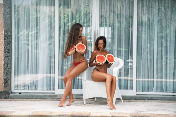 Portrait two brunette in swimsuit with watermelon fruit holding in hands and cover the chest. Sexy model with perfect body, cute smile posing and sitting on the chair. Concept of sport, vegetarian and — Stock Photo, Image