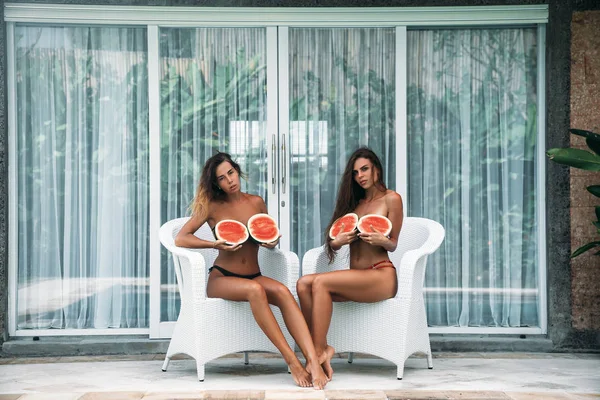 Portret dwóch brunetka w strój kąpielowy z owoców arbuza, trzymając w ręce i przykryć klatkę piersiową. Sexy model z doskonałego ciała, pozowanie uroczy uśmiech i siedzi na krześle. Pojęcie sportu, wegetariańskie i — Zdjęcie stockowe