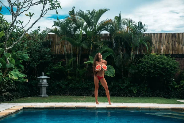 A stunning brunette girl with a sporty body holds watermelons in her hands and covers her breasts. Photoshoot of a beautiful model with fruits near swimming pool with blue clear water and green palms — Stock Photo, Image