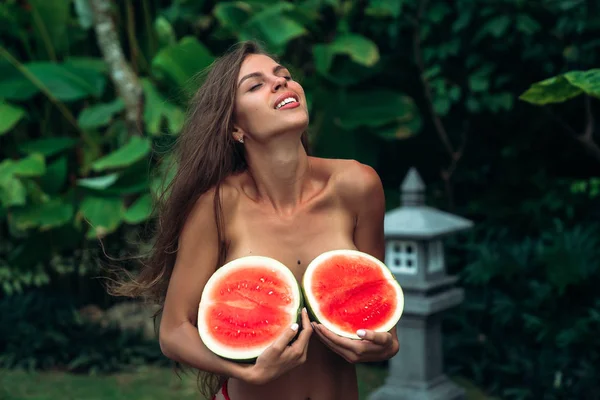 Close up portrait of cute smiling brunette girl with a sporty body holds watermelons in her hands and covers her breasts. Photoshoot of a beautiful model with fruits near green palms on background in — Stock Photo, Image