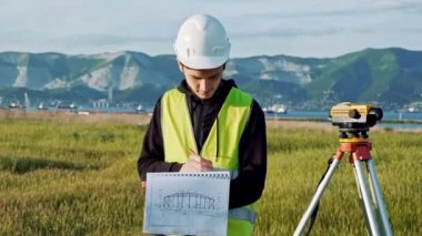 yeşil iş elbiseleri ve kask Surveyor erkekte ekipman ayarlar, hesaplamalar üretir ve inşaat alanının üstünde belgili tanımlık defter yazar. Peyzaj konsept tasarımı, Jeodezi