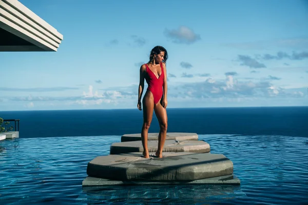Hermosa vista de chica sexy relajarse en la piscina con el océano en el fondo. Linda mujer joven en bikini rojo posando. Concepto de traje de baño, traje de baño y viajes — Foto de Stock