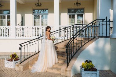 Zarif gelin düğün çiçek kokulu, gelinlik çekici kadın. Beyaz elbiseli genç kız güzel beyaz bir ev merdiven gider. Mutlu yeni evli bir kadın. Gelin düğün makyaj ile