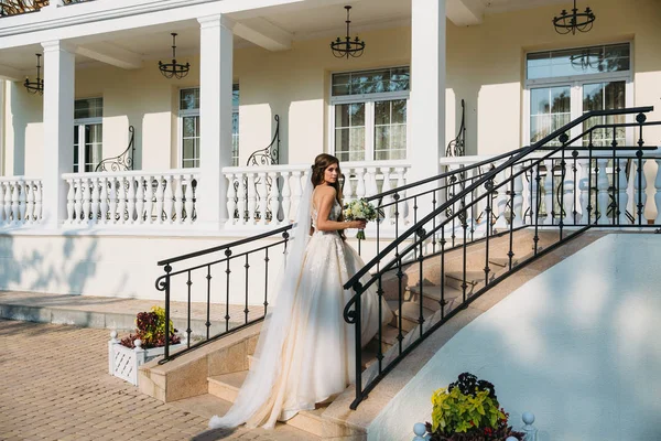 Elegante Braut mit Hochzeitsblumenstrauß, attraktive Frau im Brautkleid. junges Mädchen in weißem Kleid geht die Treppe eines schönen weißen Hauses hinauf. glückliche frisch verheiratete Frau. Braut mit Hochzeitsschminke — Stockfoto