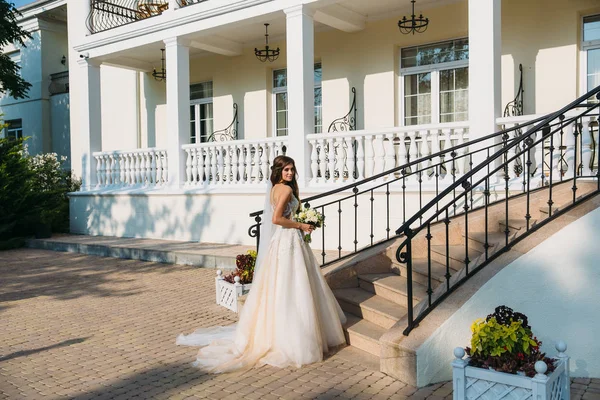 Elegante Braut mit Hochzeitsblumenstrauß, attraktive Frau im Brautkleid. junges Mädchen in weißem Kleid geht die Treppe eines schönen weißen Hauses hinauf. glückliche frisch verheiratete Frau. Braut mit Hochzeitsschminke — Stockfoto