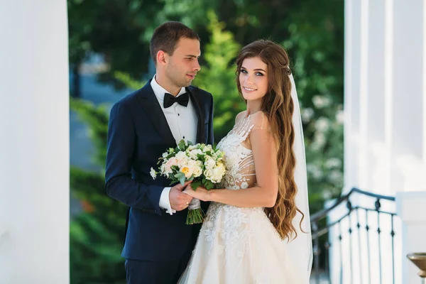 행복 한 커플 포옹과 푸른 나무와 정원에서 웃 고. 나비 넥타이와 검은 양복에 신부와 신부는 아름 다운 하얀 웨딩 드레스 그녀의 손에 꽃의 꽃다발. 결혼 — 스톡 사진