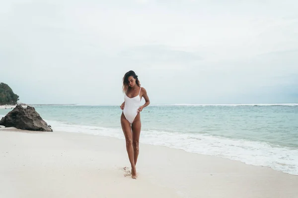 Menina incrível em um maiô branco com um belo corpo de esportes andando e posando em uma praia de areia branca. Uma jovem bronzeada com cabelo encaracolado está descansando e tomando sol. Modelo encantador posando perto do — Fotografia de Stock