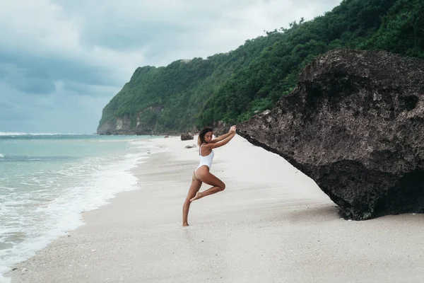Şaşırtıcı kız yürüyüş ve bir beyaz kum plajı üzerinde poz güzel spor vücut ile beyaz bir mayo. Yanık tenli genç bir kadın kıvırcık saçlı dinlenme ve güneşlenme. Yakın model poz büyüleyici — Stok fotoğraf
