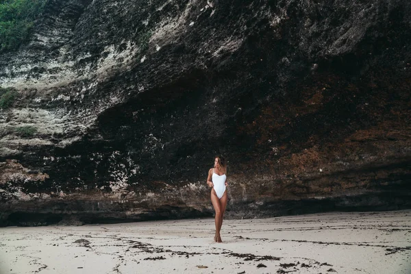 Erstaunliche Mädchen in einem weißen Badeanzug mit einem schönen Sportkörper gehen und posieren an einem weißen Sandstrand. Eine braun gebrannte junge Frau mit lockigem Haar ruht und sonnt sich. Charmantes Model posiert in der Nähe der — Stockfoto