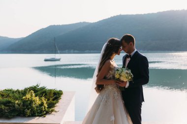 İnanılmaz evlilik çift, gelin ve damat bir göl arka plan üzerinde elele. Sevimli kız beyaz elbiseli siyah giyen adamlar uygun. Tekne ve dağlar manzara. Aile kavramı