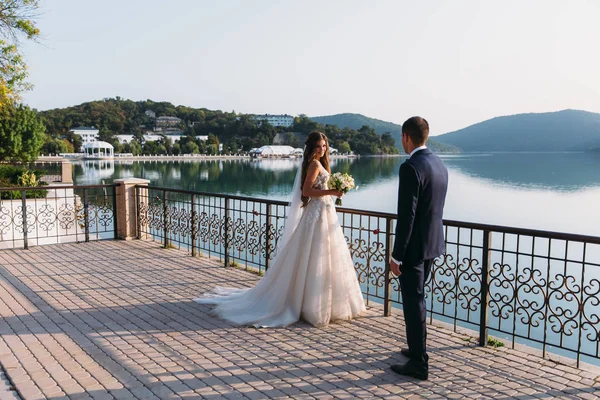 美丽的新婚夫妇, 新娘和新郎牵手在湖的背景。穿着白色连衣裙的可爱女孩, 穿着黑色西装的男人。小船和山风景。家庭概念 — 图库照片