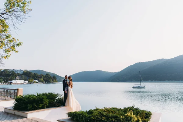 Vackra bröllopsparet, bruden och brudgummen håller händerna på sjön bakgrund. Söt flicka i vit klänning, män i svarta affärer passar. båt och berg landskap. Begreppet familj — Stockfoto