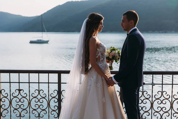 O par de recém-casados, a noiva e o noivo que mantêm mãos olharam em uns a outros olhos em um contexto de lago. Menina bonito em vestido branco, homens em terno de negócios preto. barco e montanhas paisagem. Conceito de — Fotografia de Stock