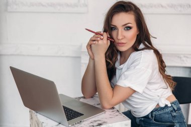 bir düşünceli freelancer kız ofiste onun elinde bir kalem ile. Rahat kıyafetler içinde bir kadın çalışıyor. Parlak bir odada bir bilgisayarla ev kadını. Yakın çekim portre.