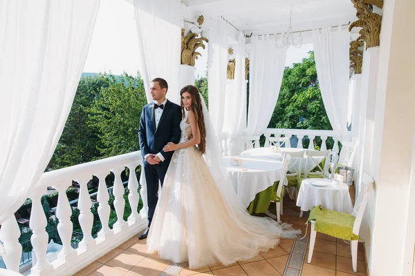 Schöne Paar Flitterwochen umarmen einander und stehen auf einem weißen Balkon. junger Bräutigam im schwarzen Anzug und eine schöne Braut mit langen Haaren im Brautkleid, die Händchen hält. — Stockfoto