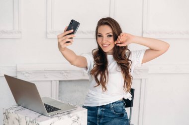 Bir dizüstü bilgisayar ve onun elinde telefon ile sürpriz kız serbest meslek selfie yapar. Beyaz bir T-shirt ve jeans Smartphone'da fotoğraflandı ve parlak bir ofiste gülümseyen genç kadınla