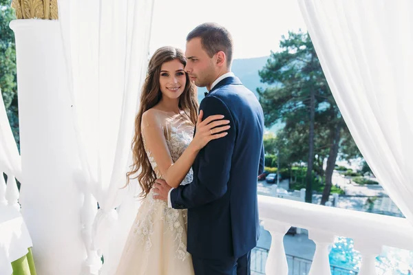 Hermosa pareja luna de miel abrazándose y de pie en un balcón blanco. Joven novio en un traje negro y una hermosa novia con el pelo largo en un vestido de novia de la mano . — Foto de Stock