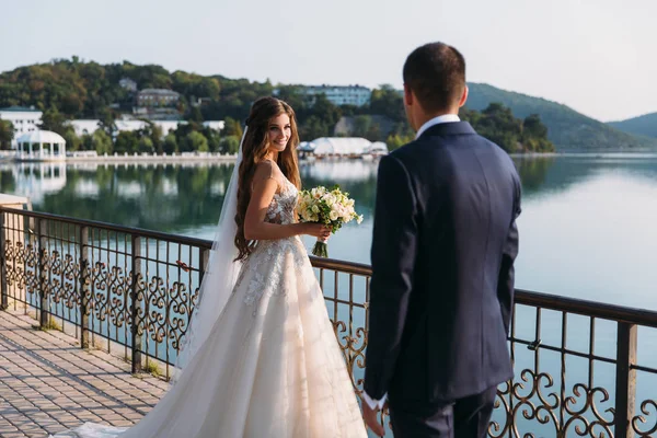 白いドレス新郎を見て、かわいい笑顔で長い髪の美しい花嫁。結婚式の日、湖の背景に魅力的なカップルの結婚式のカップル。屋外の公園で幸せな家族. — ストック写真