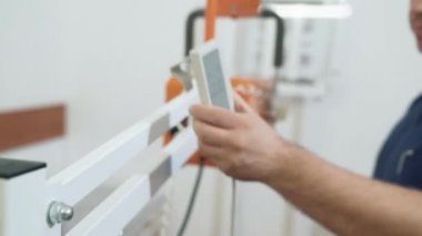 Yetişkin erkek doktor mavi üniformalı bir X-ray kontrol panelini kullanarak ayarlar. Tıbbi Araçlar Yardımcısı kurar.