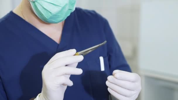 Detailní záběr lékař ve sterilní bílé rukavice a s maskou na obličeji zkoumá lékařských nástrojů v nemocnici. chirurg v profesní oblečení zkoumá svorku v rukou — Stock video