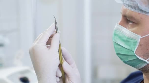 La cara cercana del médico con guantes blancos estériles y con una máscara en la cara examina los instrumentos médicos en el hospital. cirujano vestido con ropa profesional examina la abrazadera en las manos . — Vídeo de stock