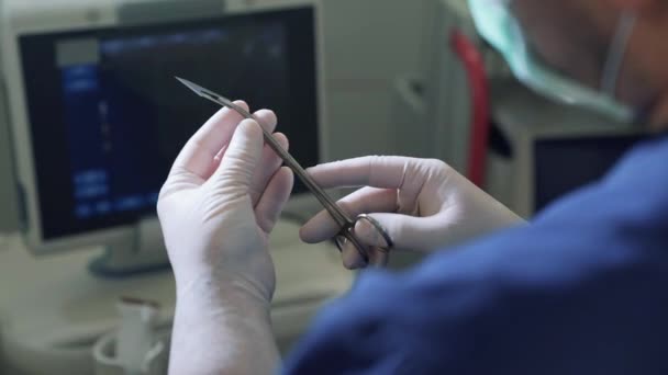 Лично доктор в стерильных перчатках держит скальпель в зажиме перед операцией. POV закрывает медицинские инструменты в руках, вид сзади, оборудование в клинике и — стоковое видео