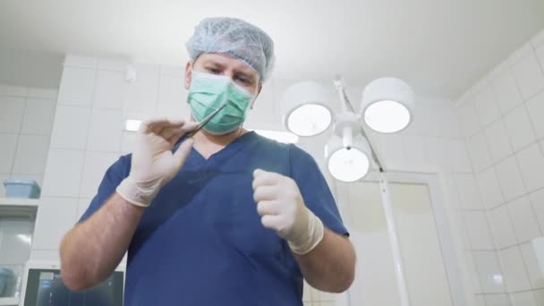 Médecin en gants blancs stériles et avec un masque sur le visage examine les instruments médicaux à l'hôpital. chirurgien dans la clinique vêtu de vêtements professionnels examine la pince dans les mains . — Video