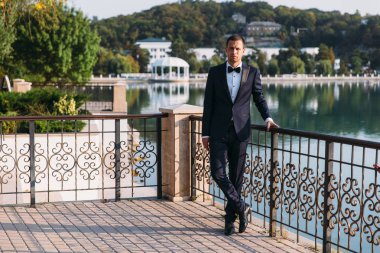 Klasik siyah bir takım elbise ile beyaz bir gömlek ve bir papyon'yetişkin erkek iş adamıdır. Parkta bir göl arka plan üzerinde bir düğün günü gelin için bekleyen damat portresi.